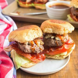 2 bacon cheeseburger sliders on round white plate with condiments in the background