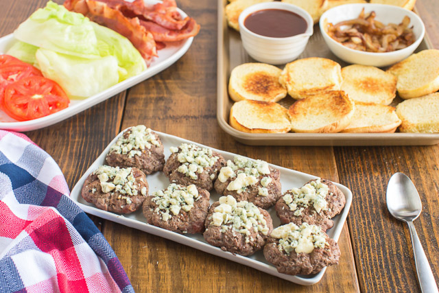 tray of 8 ground beef burgers topped with crumbled blue cheese. Trays of buns and condiments behind it