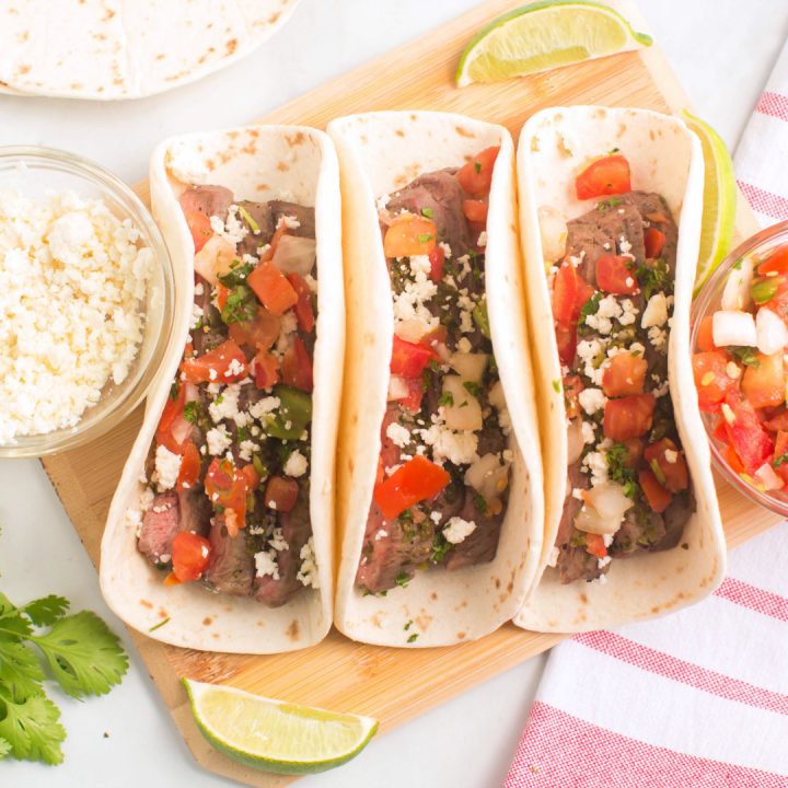 overhead photo: 3 steak street tacos on cutting board surrounded by lime wedges and Cojita cheese