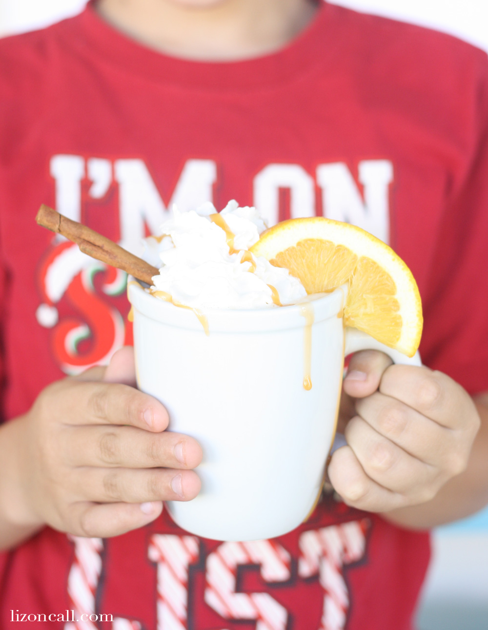 boy holding a white mug of holiday party punch, garnished with whipped cream, an orange slice, and cinnamon stick