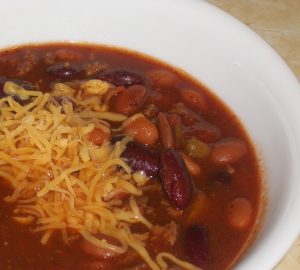 bowl of slow cooker 10 can chili topped with shredded cheddar cheese